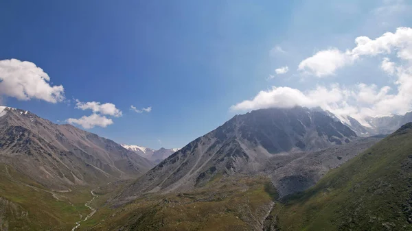 Clouds flow around the tops of high mountains. Rocky gorges and cliffs. The sky is blue, the sun is shining brightly, green grass is growing in places. Mountain terrain from the height of the drone