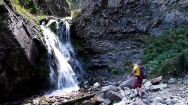 Sırt çantalı bir adam şelalenin yanında duruyor.
