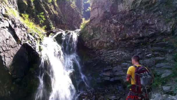 Ein Mann mit Rucksack steht in der Nähe eines Wasserfalls — Stockvideo