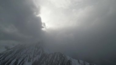 Dramatik dağ sahnesinde karlı zirvelerde yuvarlanan bulutlar, dinamik hava değişimlerini ve atmosferik derinliği gözler önüne serer, nefes kesici manzara boyunca kamera tepsileri