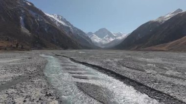 Kayalık bir vadide kıvrılan bir nehrin manzarası, arka plandaki görkemli dağları gösteriyor, kamera ileri doğru hareket ediyor, manzaranın dingin güzelliğini yakalıyor.
