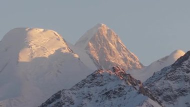Görkemli dağ zirveleri sıcak güneş ışığıyla yıkanırken, engebeli dokular ve karla kaplı yamaçlar sergilenirken, kamera manzara ve atmosferin karmaşık detaylarını gözler önüne seriyor.