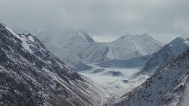 Karla kaplı görkemli dağlar bulutlu bir gökyüzüne çarpıcı bir şekilde yükseliyor, engebeli araziyi ve buzulların özelliklerini gözler önüne seriyor, kamera nefes kesici manzarayı gözler önüne seriyor.