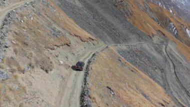 Bir araç engebeli, dolambaçlı toprak bir yolda ustalıkla dolaşır, doğal manzara dokularını ve renklerini gösterir, dinamik hava kamerası hareketleriyle sahneyi güçlendirir.