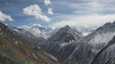 Görkemli dağ manzarası karla kaplı tepeleri ve kıvrımlı bir nehri gösteriyor. Kamera nefes kesen manzarayı yansıtıyor. Kayalık arazinin dokularını ve canlı gökyüzünü gözler önüne seriyor.
