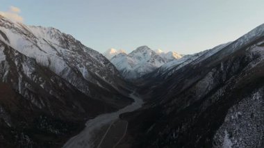 Hava görüntüsü, karla kaplı dağları ve aşağıdaki geniş vadiyi yakalar, doğal güzelliği ve engebeli araziyi gözler önüne serer, kamera peyzaj boyunca düzgün bir şekilde hareket eder.