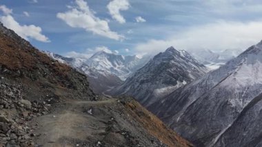 Kayalık arazide dolambaçlı bir yol sunan görkemli dağ manzarası, karla kaplı tepeler ve dramatik gökyüzü, kamera nefes kesici manzaralar boyunca hareket ediyor.