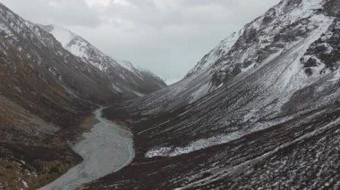 Görkemli dağ vadisinde, karlı tepeler ve kayalık arazilerle çevrili dolambaçlı bir nehir bulunur. Kamera nefes kesen manzarayı ve doğal güzelliği sorunsuzca yansıtır.