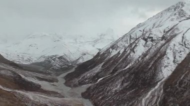 Karlı dağ manzarası derin bir vadiden akan kıvrımlı bir nehri, engebeli araziyi ve dramatik tepeleri gösterir, kamera geniş manzarayı ortaya çıkarır.