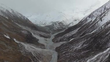 Hava perspektifi, karlı dağları yakalar engebeli bir vadiden akan kıvrımlı bir nehri çerçeveler, dramatik manzarayı ve atmosferik koşulları gözler önüne serer, kamera yavaşça uzaklaşır.