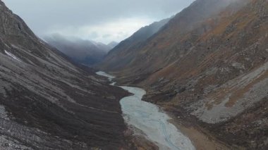 Hava görüntüleri engebeli bir dağ vadisinde akan kıvrımlı bir nehri yakalıyor, kamera sahnenin derinliğini ve güzelliğini ortaya çıkarmak için pürüzsüz bir şekilde hareket ederken dramatik manzarayı gösteriyor.
