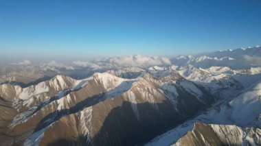 Kar kaplı bir dağ silsilesinin nefes kesici manzarası ufuk boyunca uzanıyor, yukarıdaki açık mavi gökyüzü ve yumuşak bulutlarla kısmen örtülü uzak zirveler doğal güzellikleri gösteriyor.