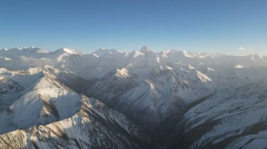 Ufuk boyunca uzanan karla kaplı bir sıradağdan nefes kesici bir manzara, açık mavi gökyüzünün altında dramatik tepeler ve vadiler sergiliyor, doğanın güzelliğini vurguluyor.