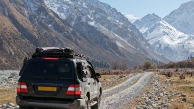Bir off-road aracı heybetli dağlar ve berrak mavi gökyüzü ile çevrili kayalık bir yolda ilerliyor, engebeli arazileri ve güzellikleri keşfetme macerasını gözler önüne seriyor.