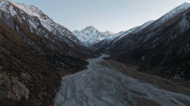 Karla kaplı zirveler dolambaçlı bir nehir vadisinde dramatik bir şekilde yükselir, doğanın engebeli güzelliğini gözler önüne serer. Sakin manzarada kayalık arazi ve yemyeşil bitki örtüsü nefes kesici bir manzara yaratıyor.