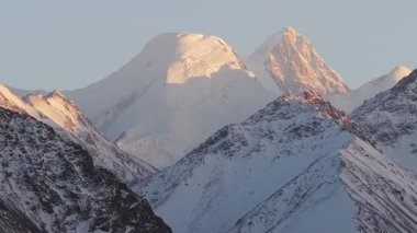 Görkemli kar örtülü dağlar altın güneş ışığıyla yıkanır, engebeli dokular ve dramatik tepeler sergilenir, kamera düzgünce yakınlaşır ve manzaranın karmaşık detaylarını gözler önüne serer.
