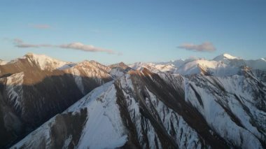 Görkemli bir dağ sırasının nefes kesici hava manzarası, karla kaplı tepeleri ve engebeli araziyi gösteriyor, kamera pürüssüz bir şekilde manzarayı tarıyor, büyüleyici doğal güzelliği gözler önüne seriyor.