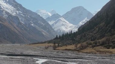 Akan bir nehir, yemyeşil yemyeşil ve yükselen zirveler içeren görkemli bir dağ manzarası. Kamera, doğanın dingin güzelliğini yansıtıyor.