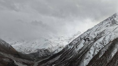Görkemli karlı dağlar bulutlu bir gökyüzü altında belirgin bir şekilde yükselirler ve aşağıdaki derin vadiyi gösterirler. Engebeli arazi ve zıt dokular nefes kesici bir doğal manzara yaratır.