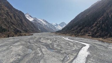 Dağlık bir arazide süzülen kayalık bir nehir yatağının geniş bir manzarası. Karla kaplı tepeler ve yemyeşil tepelerle çevrili. Doğanın ve sükunetin güzelliğini gözler önüne seriyor.