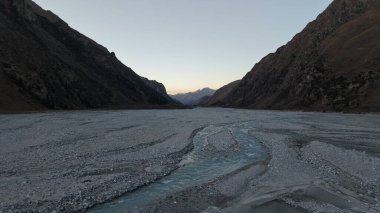 Sakin bir nehir, karanlık bir gökyüzünün altındaki görkemli dağlarla çevrili kayalık bir vadiden akar. Manzara, akşam ışığını yansıtan taş ve su dokularıyla doğanın güzelliğini sergiliyor.