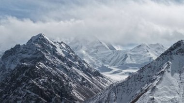 Karla kaplı görkemli dağ zirveleri bulutlu bir gökyüzüne çarpıcı bir şekilde yükselir, doğanın engebeli güzelliğini ve yüksek irtifadaki manzaranın sakin atmosferini gözler önüne serer.