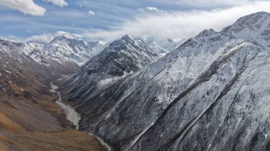 Derin bir vadideki kıvrımlı bir nehrin üzerinde yükselen karla kaplı dağ zirveleri kayalık arazi ve dramatik bulut oluşumlarıyla doğanın güzelliğini gözler önüne seriyor.