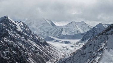 Karla kaplı sıradağlar bulutlu bir gökyüzünün altında görkemli bir şekilde yükselirler dramatik tepeler ve vadiler sergilerler, nefes kesen doğal bir manzara yaratarak macera ve sükunet hissi uyandırır.
