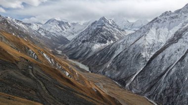 Dolambaçlı bir nehir vadisini çevreleyen karla kaplı dağların nefes kesici manzarası kayalık arazi ve bulutlu bir gökyüzünün altındaki altın çimen parçalarıyla doğanın güzelliğini gözler önüne seriyor.