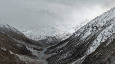 Karlı dağ vadisinin geniş bir manzarası, engebeli tepeler ve aşağıda dolambaçlı bir nehir, yoğun bulutlarla dolu dramatik bir gökyüzü altında, sakin ama güçlü bir atmosfer yaratıyor.