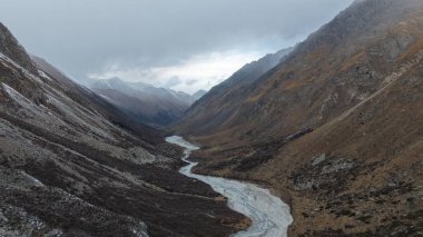 Hava perspektifi engebeli bir dağ vadisinde akan dolambaçlı bir nehri yakalar kayalık araziler ve bulutlu bir gökyüzü altında seyrek bitki örtüsü sergiler, doğanın güzelliğini ön plana çıkarır.