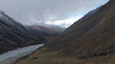 Dolambaçlı bir nehir, kayalık yamaçlar ve çimenlerin yer aldığı geniş bir dağ vadisi manzarası. Sahne, doğanın ham güzelliğini dramatik ışıklandırma ve dokularla yakalıyor.