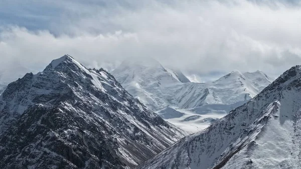 Karla kaplı görkemli dağ zirveleri bulutlu bir gökyüzüne çarpıcı bir şekilde yükselir, doğanın engebeli güzelliğini ve yüksek irtifadaki manzaranın sakin atmosferini gözler önüne serer.