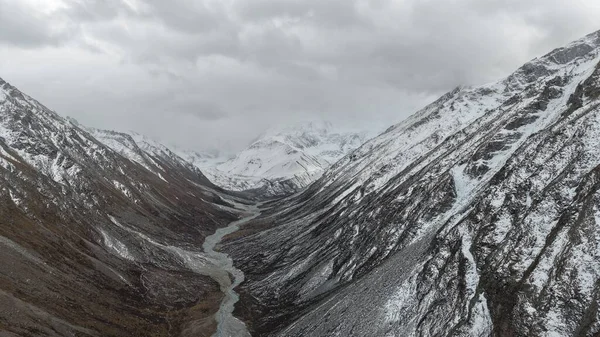 Karlı bir dağ vadisinde nefes kesici bir manzara, sarmal bir nehir manzarayı kesiyor, sarp, engebeli zirvelerle çevrili dramatik bulutlu bir gökyüzü altında, doğal güzellikler sergiliyor.