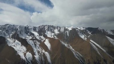 Hava manzarası, karla kaplı zirveler ve dramatik bulutlarla çarpıcı sıradağları yakalar, engebeli araziyi ve doğal güzelliği gözler önüne serer, kamera sahneyi pürüzsüz bir şekilde geçer.