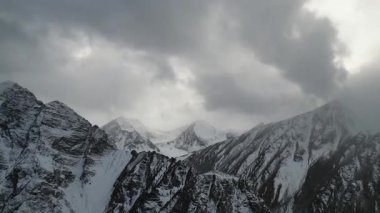 Karla kaplı görkemli dağlar bulutlarla dolu dramatik bir gökyüzü altında yükseliyor, engebeli arazi ve dokuları gözler önüne seriyor, kamera nefes kesici manzarayı tarıyor.