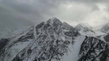 Gökyüzü görüntüsü, engebeli dokuları ve dramatik bulut oluşumlarını gösteren görkemli kar örtülü sıradağları yakalar. Kamera geniş araziyi düzgün bir şekilde gözler önüne serer.