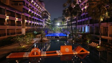 Geceleri lüks bir otel havuzunda yüzen turuncu şişme engelli parkur. Çevredeki binalar ve palmiye ağaçları tarafından aydınlatılıyor. Canlı ve davetkar bir atmosfer yaratıyor.