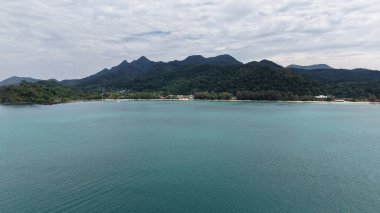 Turkuaz su, tropikal bir adanın kumlu sahilinde yavaşça dolaşır, yemyeşil dağlar bulutlu bir gökyüzünün altında ihtişamlı bir şekilde yükselir, sakin ve resimsel bir manzara yaratır.