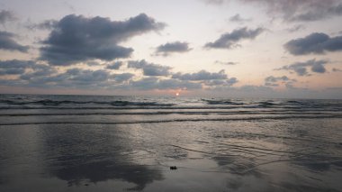 Yumuşak dalgalar yavaşça kumlu kıyıya vuruyor güneş ufkun altına batarken, plajda sakin bir günbatımında, turuncu ve pembe renkli canlı gökyüzünü boyuyor.