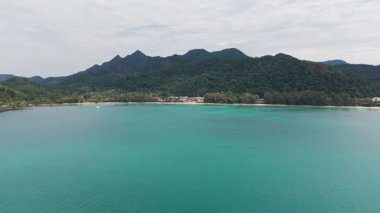 Hava perspektifi turkuaz sulardan yükselen yeşil dağlık adayı yakalıyor, bulutlu gökyüzünün altında sakin tropikal manzarayla el değmemiş kıyı şeridini ortaya çıkarıyor.