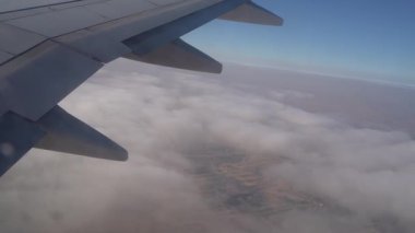 Bulutların üzerinde uçarken uçak kanatları görünür, dağınık yapılar ve yollar ile yavaş yavaş geniş bir çöl manzarasını ortaya çıkarıyor, gökyüzünden yere geçişi gösteriyor.