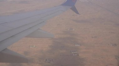 Uçak kanadı geniş bir çöl arazisine yayılarak yerdeki dağınık yapıları ortaya çıkarır. Uçak inerken görüntüler kurak araziyi ve uzak şehir manzarasını gösteriyor.