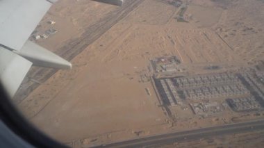 Bir uçağın hava perspektifi dağınık binalar ve bir yol ile geniş bir çöl manzarası gösteriyor, kentsel yapılar ve çevre kumlu arazi arasındaki zıtlığı vurguluyor.