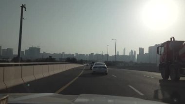 Aralarında beyaz bir sedan ve çimento karıştırıcı kamyonun da bulunduğu araçlar, işlek bir yol sahnesini gözler önüne seren, parlak güneş ışığı altında şehrin ufuk çizgisini net bir şekilde gören şehir otoyolu boyunca ilerliyorlar.