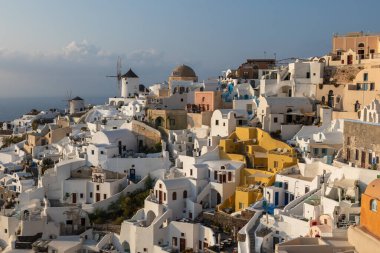 Oia, Santorini Adası, Yunanistan - 18 Eylül 2020: Köydeki geleneksel siklodik beyaz mimarinin manzaralı manzarası. Arka planda ünlü yel değirmenleri. Günbatımı yaşam noktası.