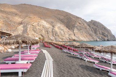 Yunanistan 'ın Santorini adasındaki Perissa plajının üst manzarasında pembe güneşlikler ve şemsiyeler var. Plaj siyah, volkanik kumlarla kaplı, Yunanistan.