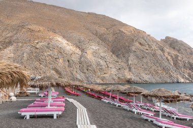 Yunanistan 'ın Santorini adasındaki Perissa plajının üst manzarasında pembe güneşlikler ve şemsiyeler var. Plaj siyah, volkanik kumlarla kaplı, Yunanistan.