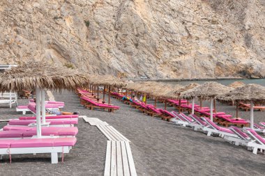 Yunanistan 'ın Santorini adasındaki Perissa plajının üst manzarasında pembe güneşlikler ve şemsiyeler var. Plaj siyah, volkanik kumlarla kaplı, Yunanistan.