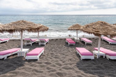 Yunanistan 'ın Santorini adasındaki Perissa plajının üst manzarasında pembe güneşlikler ve şemsiyeler var. Plaj siyah, volkanik kumlarla kaplı, Yunanistan.